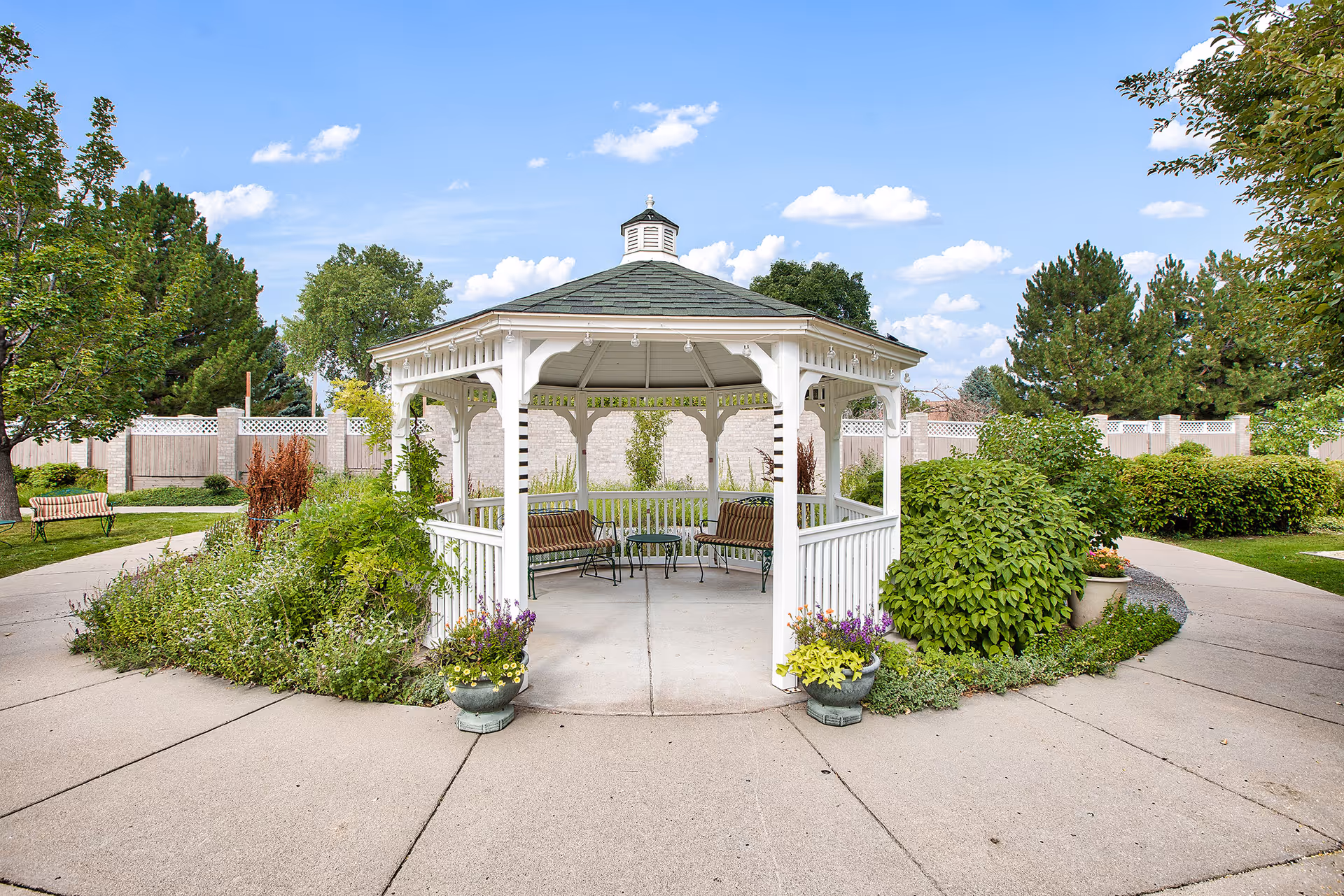 A white wooden gazebo with a green roof situated in a garden area. The gazebo has benches and a small table inside. Surrounding the gazebo are paved walkways, green bushes, trees, and flower pots with colorful flowers. The sky is blue with scattered clouds.