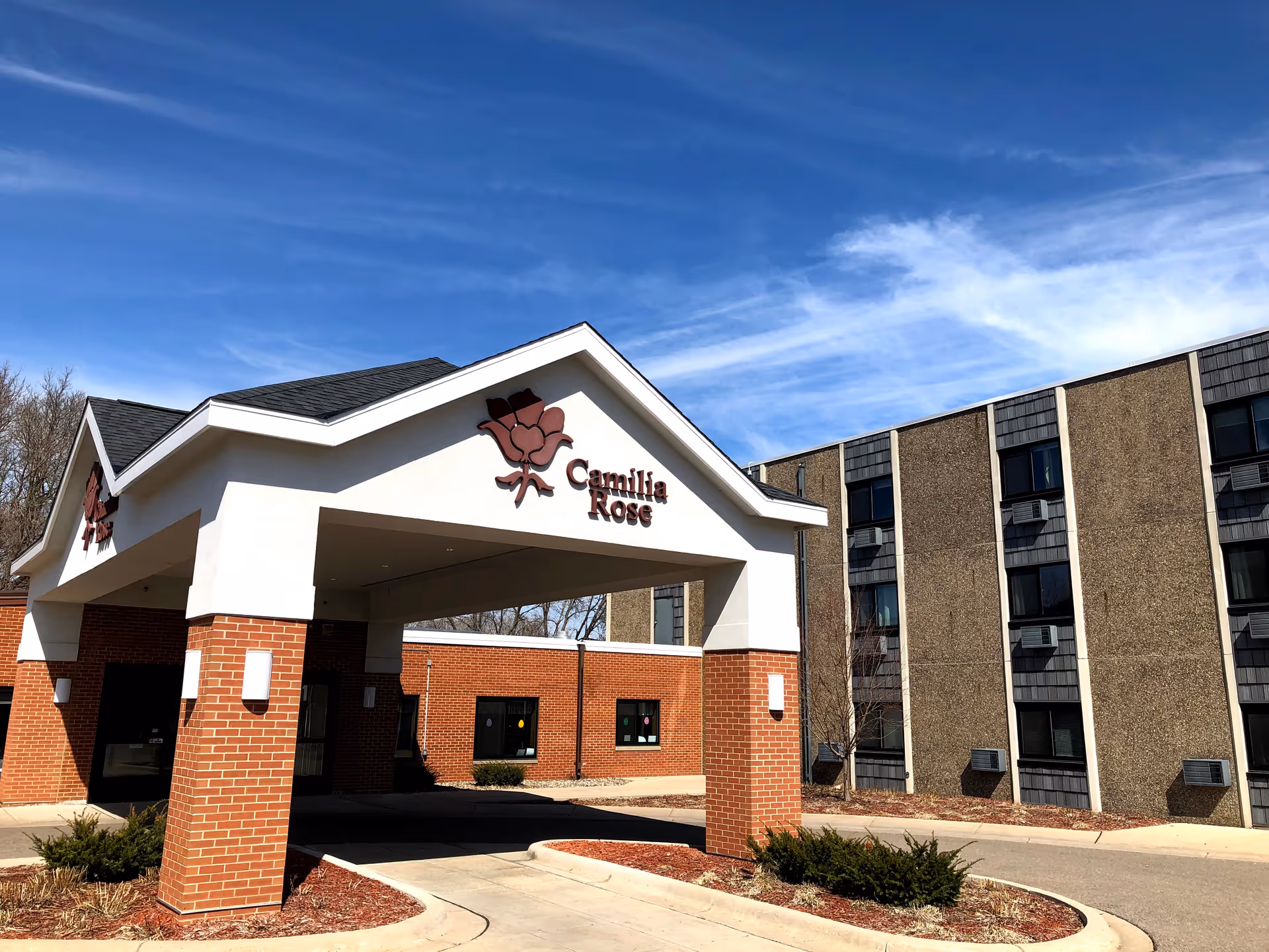 Exterior view of Camilia Rose Care Center showing the entrance canopy with a rose logo and the facility name. The building has a brick and beige facade with multiple windows and air conditioning units. The sky is clear with some wispy clouds.