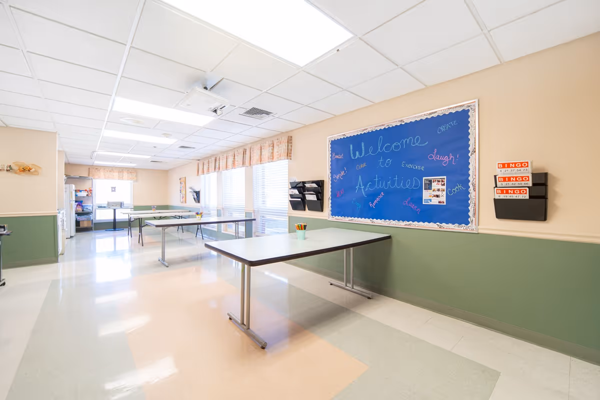Bright activity room with folding tables, a blue 'Welcome to Activities' bulletin board, bingo cards on the wall, and large windows.