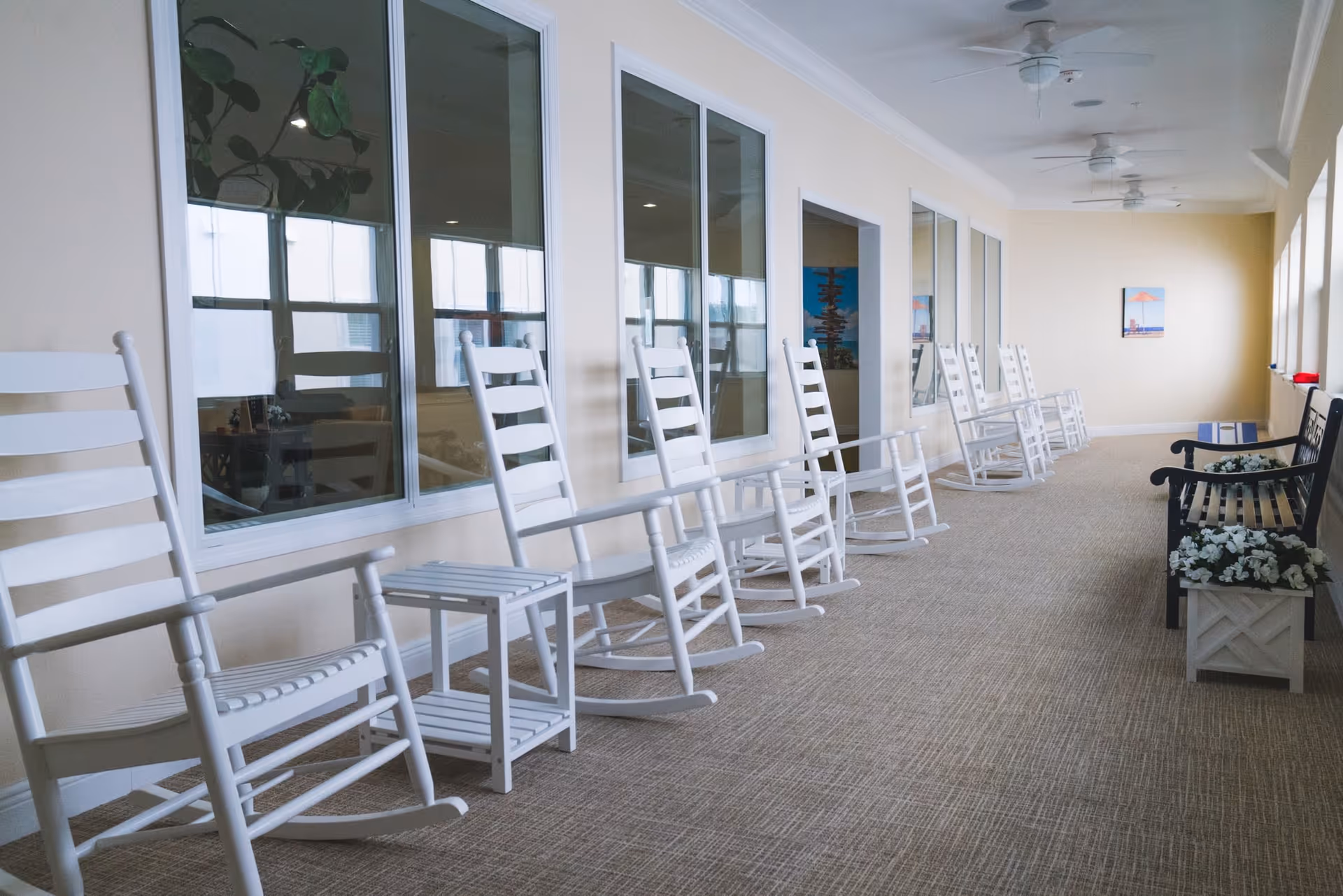 A long indoor hallway with a row of white wooden rocking chairs and small tables along one wall, large windows on the opposite wall, and a bench with flower boxes at the far end.