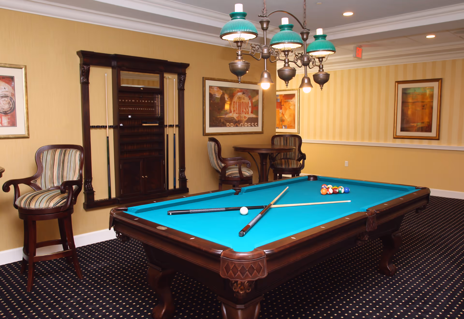 A well-lit game room featuring a pool table with a teal felt surface, two pool cues resting on the table, and a rack with additional cues mounted on the wall. The room has patterned carpet, striped upholstered chairs, framed artwork on yellow striped walls, and a vintage-style chandelier with green shades hanging above the pool table.