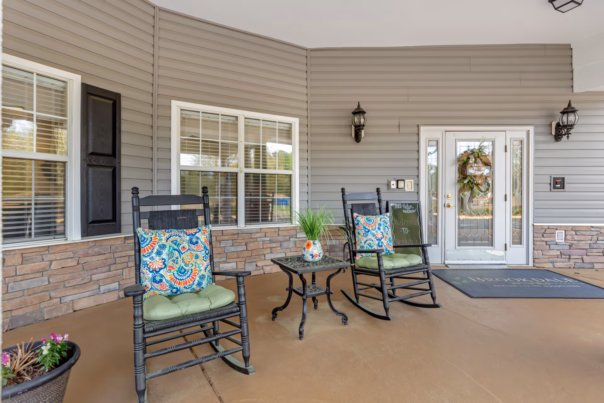 Covered porch area with two black rocking chairs featuring colorful patterned cushions and green seat pads, a small black metal table with a decorative vase holding green plants, a welcome mat with the Brookdale logo, a door with glass panels and a wreath, and a chalkboard sign welcoming visitors to Brookdale.