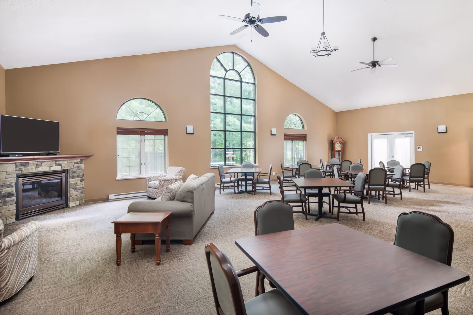 A spacious common area with high vaulted ceilings, large arched windows letting in natural light, multiple tables and chairs arranged for group seating, a stone fireplace with a TV mounted above it, and comfortable sofas and armchairs.