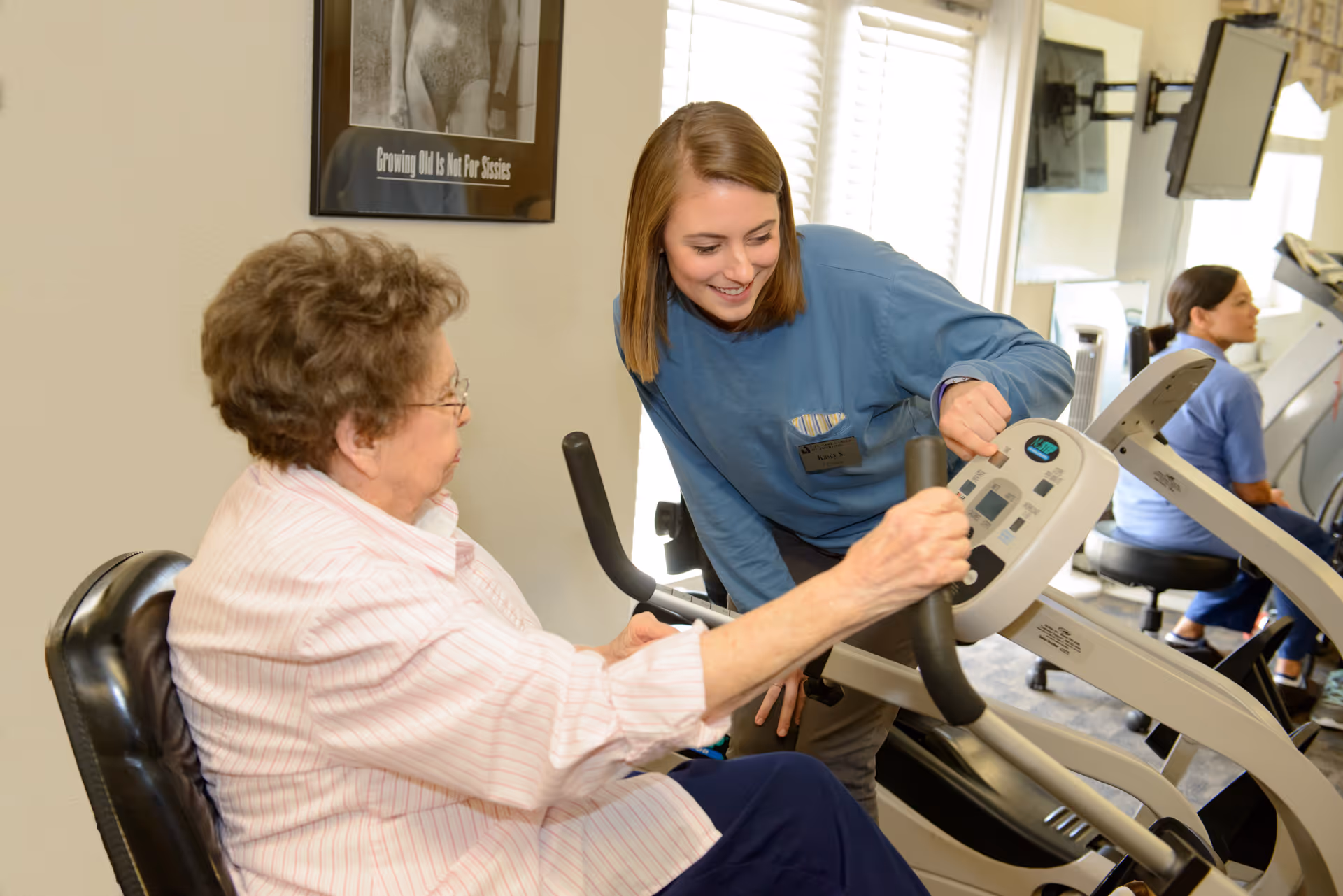 An elderly woman using a stationary exercise bike with the assistance of a smiling female caregiver in a fitness room. Another person is visible in the background using exercise equipment. The room has large windows letting in natural light and a framed picture on the wall.