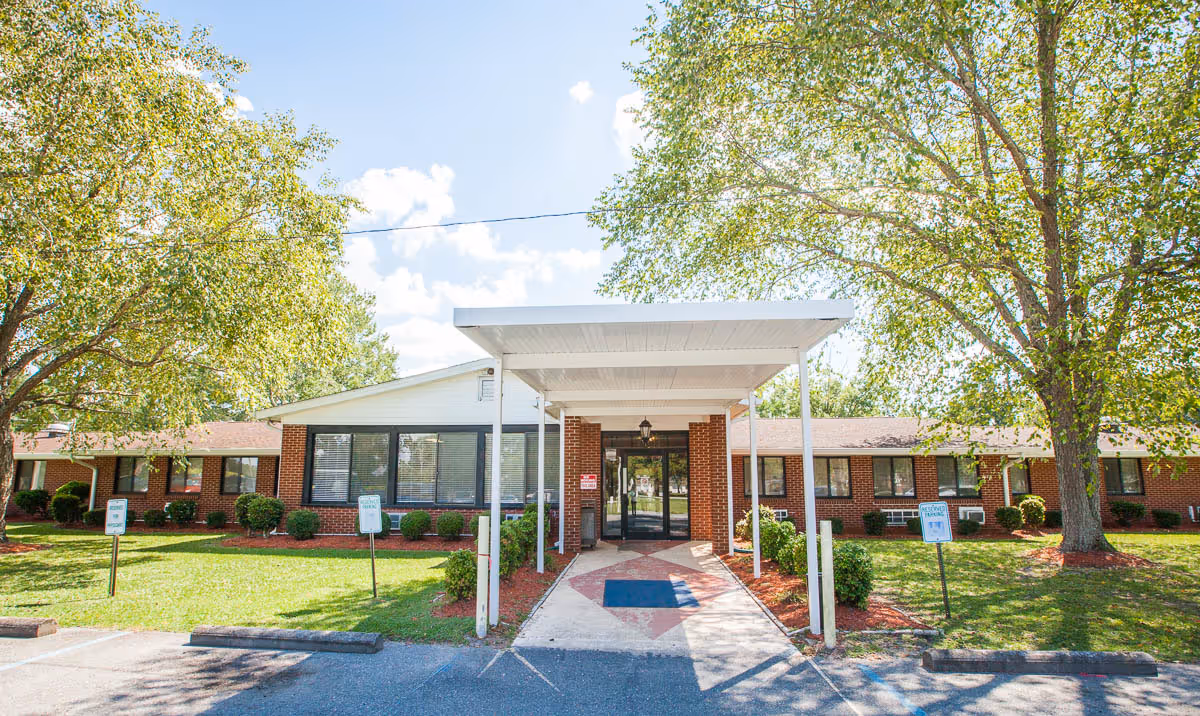 Front exterior view of a single-story brick building with large windows, a covered entrance with white pillars, surrounded by green grass and trees under a blue sky with some clouds.
