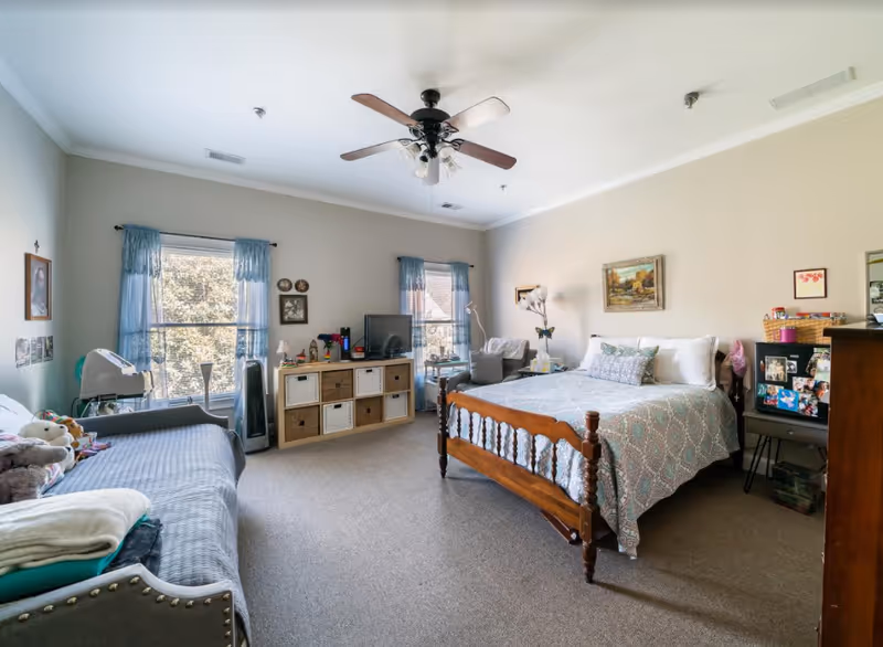 A spacious bedroom with a wooden bed covered in a patterned bedspread, a gray couch with stuffed animals, two windows with blue curtains, a ceiling fan, a TV on a storage unit with baskets, and various framed pictures on the walls.