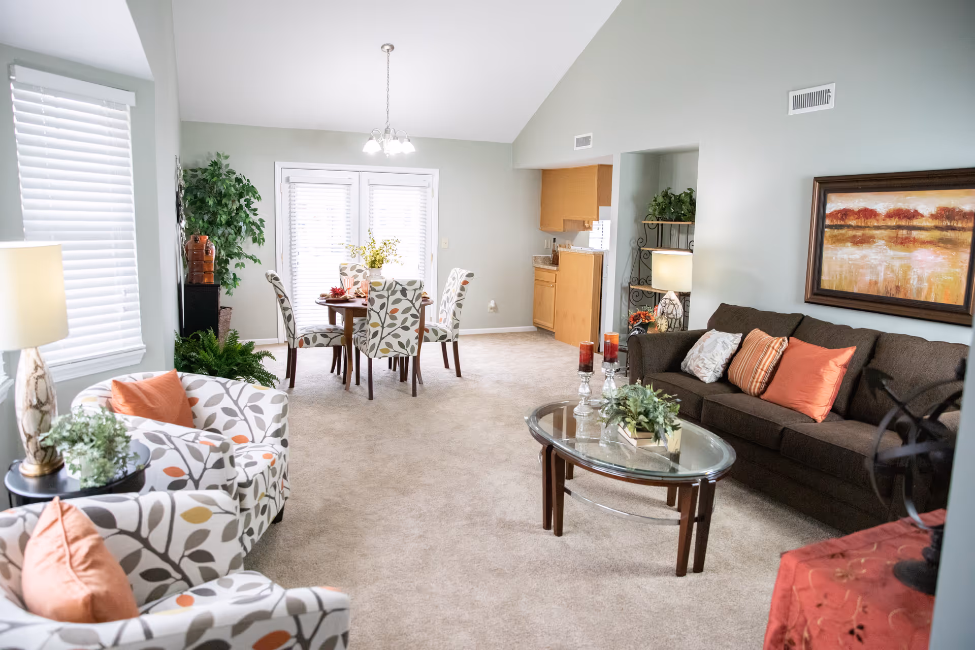 Bright senior living room with patterned chairs, brown couch, glass coffee table, and dining area.