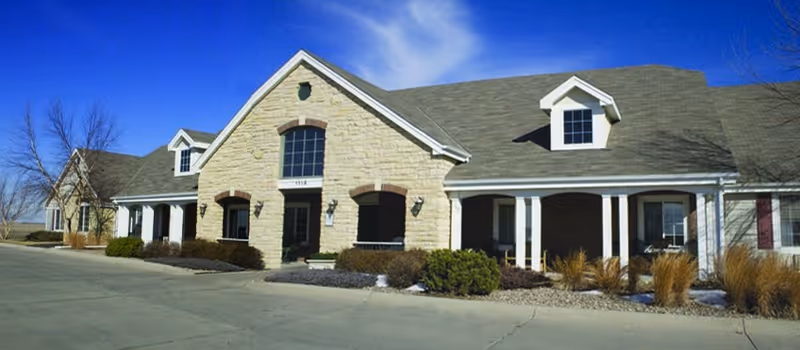 Exterior view of a single-story assisted living facility building with beige stone facade, multiple windows, a covered porch with white columns, and a gray shingled roof under a clear blue sky.