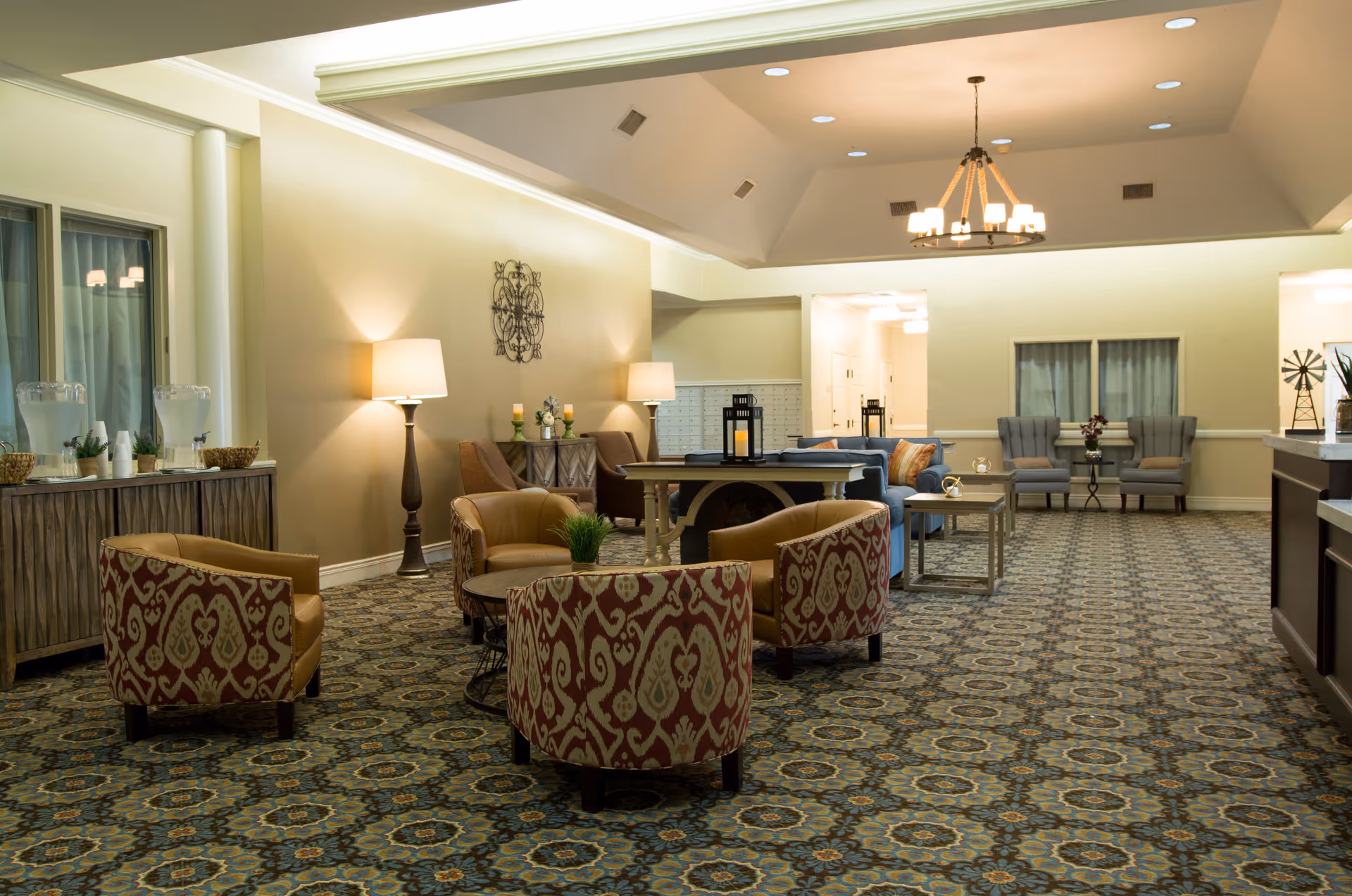 A spacious and well-lit common lounge area with patterned carpet, several armchairs with red and beige upholstery, a round table with a small plant, two floor lamps, a blue sofa with colorful cushions, and additional seating including two gray armchairs near a window. The room has a high ceiling with recessed lighting and a chandelier, and a sideboard with water dispensers and cups.