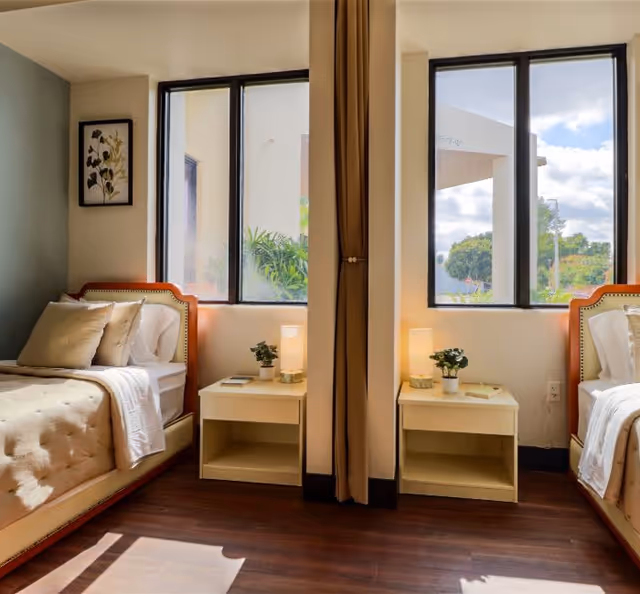 A bright and cozy bedroom with two single beds separated by a curtain. Each bed has beige bedding and pillows. Between the beds are two small nightstands, each with a small potted plant, a book, and a lit lamp. Large windows behind the beds let in natural light and show greenery outside. A framed botanical print hangs on the wall to the left.