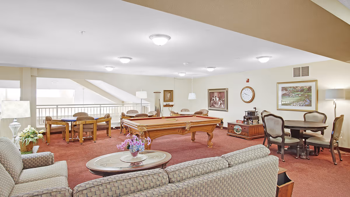 A spacious common area in a senior living facility featuring a pool table at the center, surrounded by several seating arrangements including armchairs, sofas, and tables with chairs. The room has beige walls adorned with framed artwork and a clock, red carpet flooring, and multiple ceiling lights providing bright illumination.