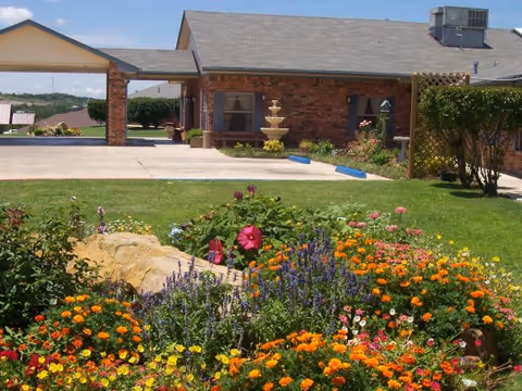 A colorful garden with various blooming flowers including orange, yellow, purple, and pink blossoms in front of a brick building with a gray roof. There is a concrete driveway and a three-tiered fountain near the building entrance. The sky is clear and blue.