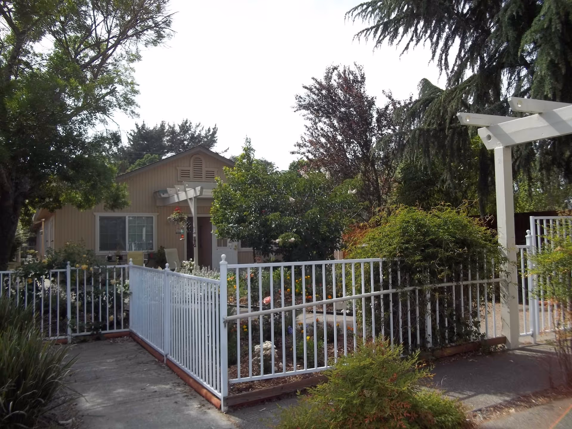 View of a single-story beige building surrounded by a white metal fence and lush greenery, including trees and bushes, under a bright sky.