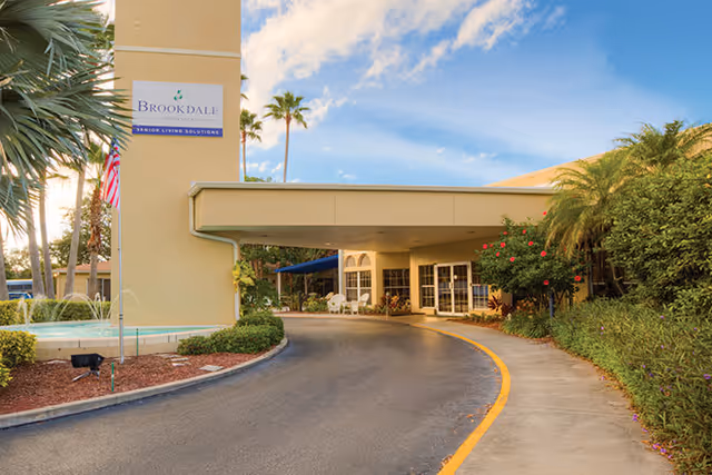 Entrance driveway and covered drop-off area of Brookdale Palma Sola senior living facility with palm trees, landscaping, and a clear blue sky.