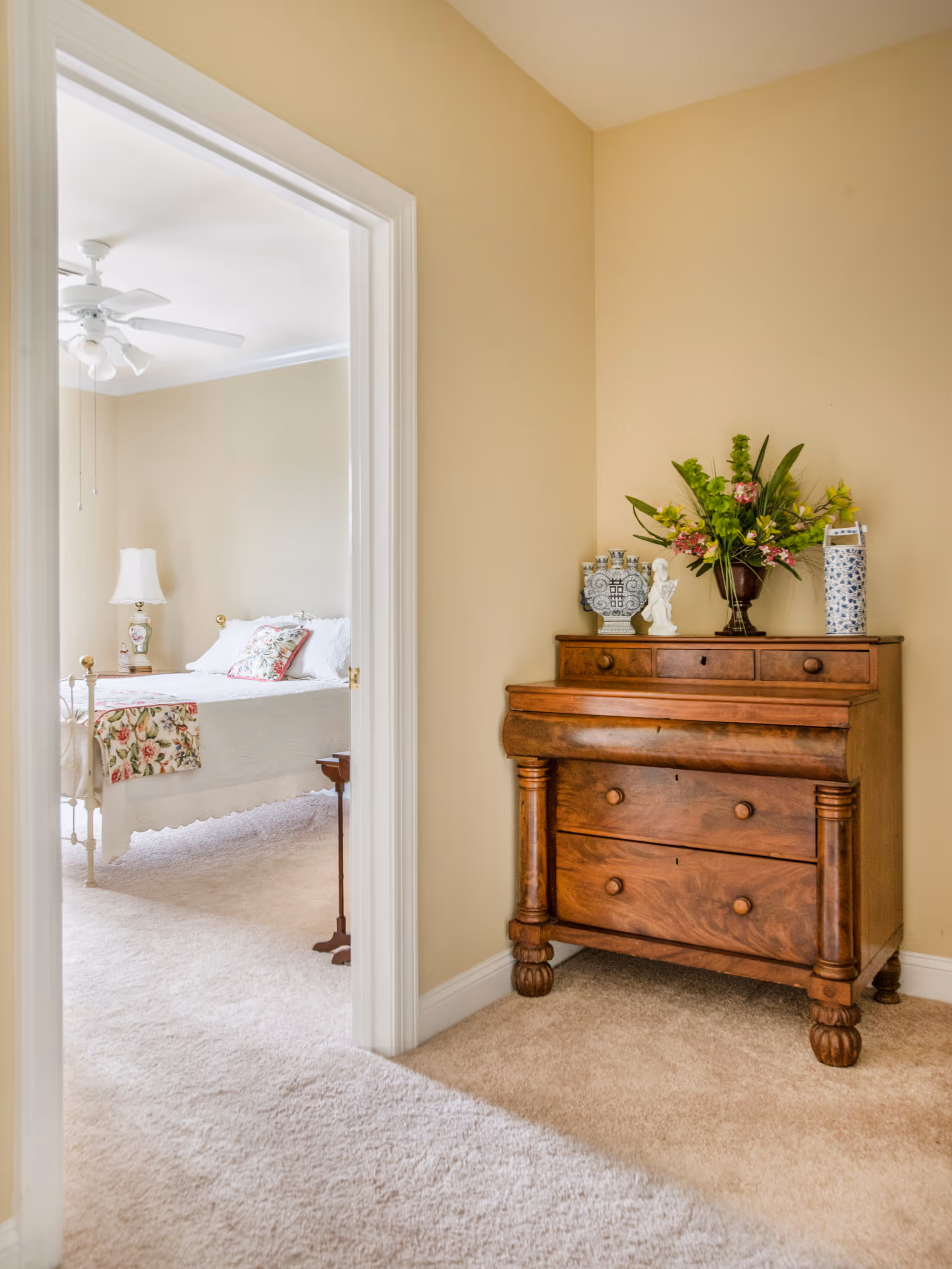 View of a cozy bedroom with a white bedspread and floral pillows, seen through an open doorway from a carpeted hallway. To the right in the hallway, there is a wooden chest of drawers decorated with a vase of flowers, a small statue, and other decorative items. The walls are painted beige and the room has a ceiling fan with lights.