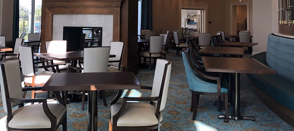 A dining area with multiple tables and chairs arranged neatly. The room features a fireplace with a wooden mantel, patterned carpet flooring, and large windows letting in natural light. The chairs are upholstered in light fabric, and there is a curved booth seating area with teal cushions on the right side.