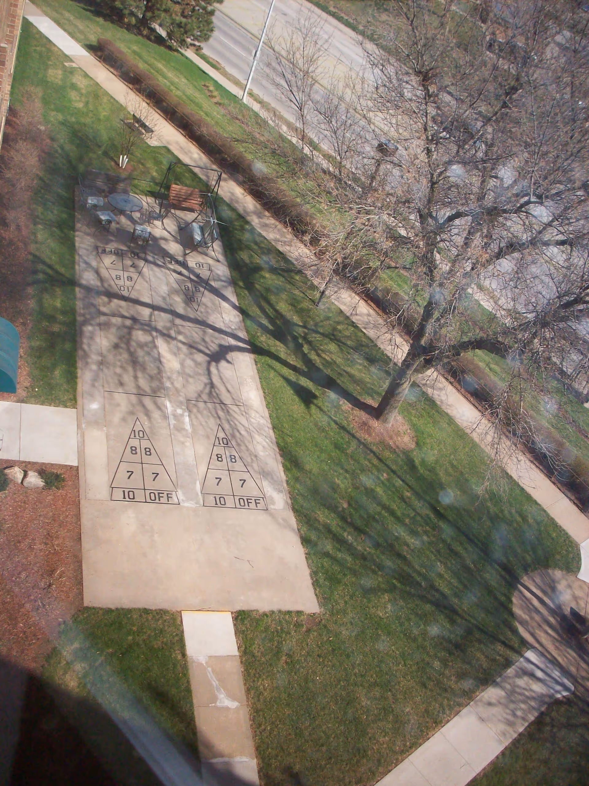 Aerial view of an outdoor shuffleboard court with two playing areas, surrounded by green grass, leafless trees, and paved walkways. There is a bench and a small table with chairs near the shuffleboard court. A road and sidewalk are visible in the background.