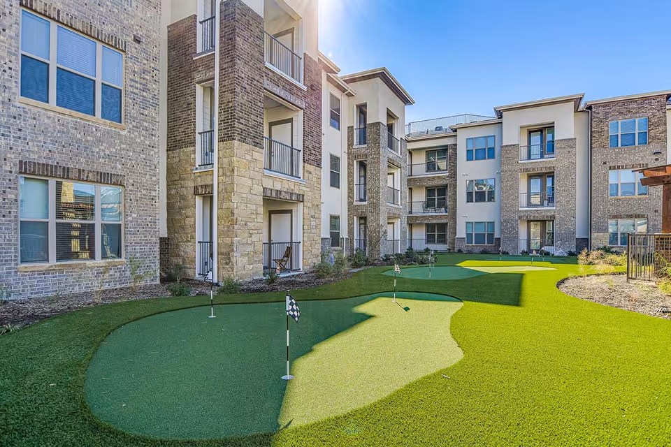 Outdoor courtyard area of a senior living facility featuring a putting green with several golf holes and flags, surrounded by a three-story brick and stone building with balconies and windows under a clear blue sky.