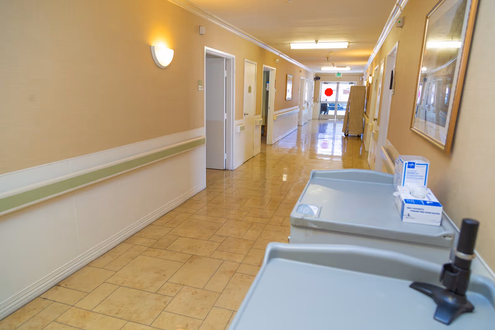 A clean, well-lit hallway in a senior living facility with beige walls, tiled floor, handrails along the walls, and several doors leading to rooms. Two medical supply carts are positioned on the right side of the hallway, and framed artwork is hung on the wall. The hallway extends toward a glass exit door at the far end.