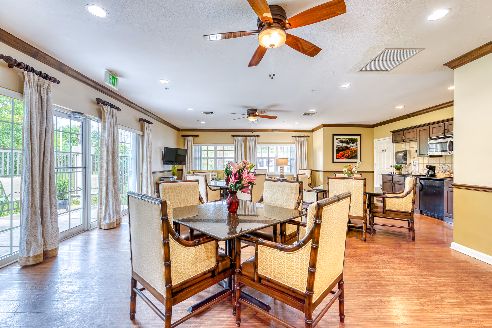Bright communal dining and lounge area with multiple tables and chairs, ceiling fans, large windows and a kitchenette.