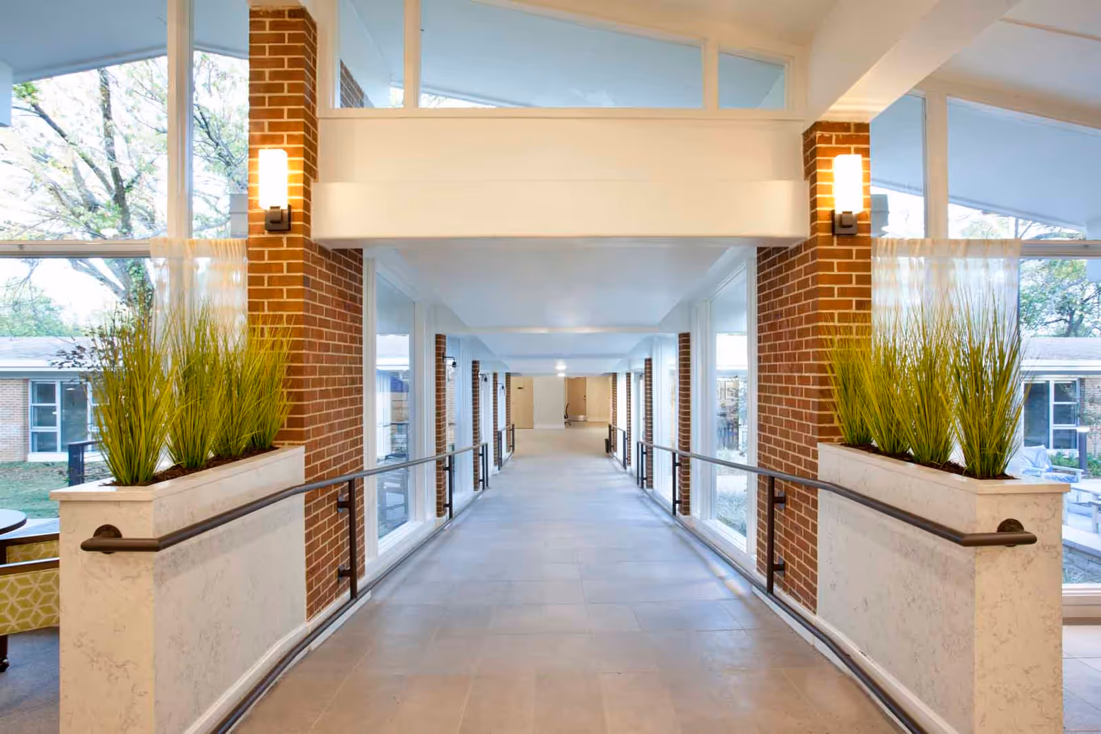 A bright indoor hallway with large windows on both sides, brick columns, and planters with tall green plants. The hallway has handrails on both sides and leads to a distant door.