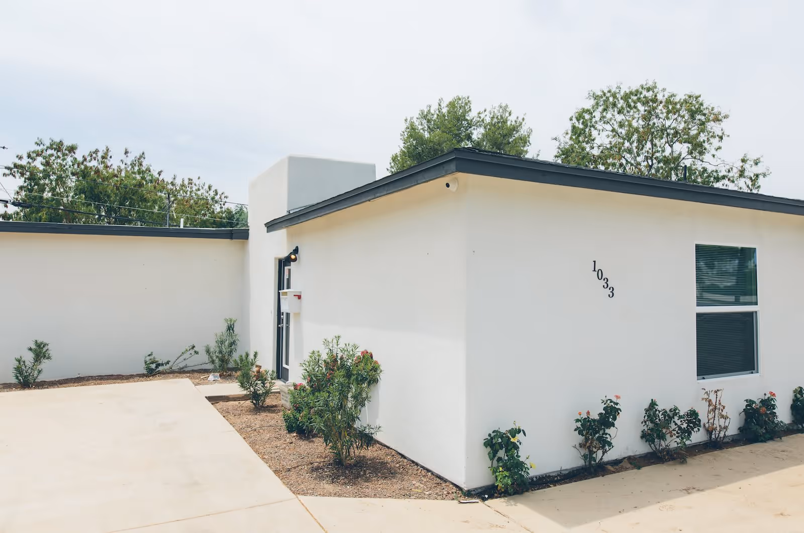 Exterior view of a modern, single-story white building with a flat roof and a small garden area with shrubs and plants along the walls. The building has a window with blinds and the number 1033 displayed on the wall. The sky is overcast and there are trees in the background.
