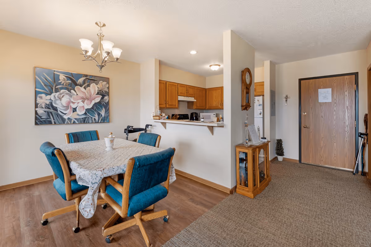 Interior view of a senior living facility showing a dining area with a table covered by a floral tablecloth and four blue cushioned chairs on wheels. A large floral painting hangs on the wall above the table. The dining area is adjacent to a small kitchen with wooden cabinets and white appliances. To the right, there is an entryway with a wooden door, a small wooden cabinet with glass doors, a wall clock, and a decorative cross on the wall. The floor transitions from wood in the dining area to carpet in the entryway.