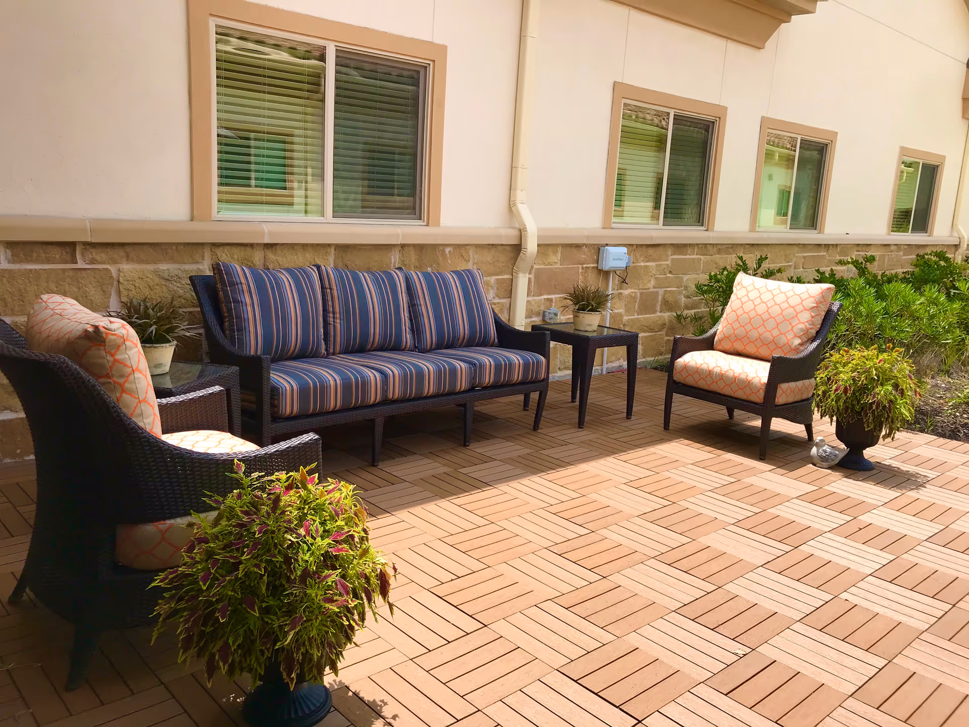 Outdoor patio area with a striped cushioned sofa, two cushioned armchairs with patterned pillows, two small side tables with potted plants, and additional potted plants on a wooden tile floor. The patio is adjacent to a building with beige walls and several windows with blinds.
