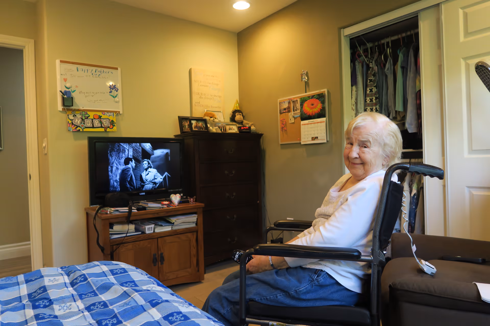 An elderly woman sitting in a wheelchair in a cozy bedroom. The room has a bed with a blue checkered blanket, a wooden dresser with framed photos and decorations, a TV showing a black and white movie, and an open closet with hanging clothes. The walls are decorated with a whiteboard, a calendar, and a motivational sign.