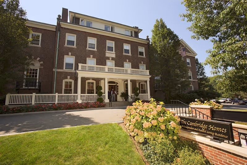 Exterior view of a large brick building with multiple windows and a white porch entrance. There is a well-maintained lawn and flower bushes in front, along with a sign that reads 'The Cambridge Homes'. Trees and a clear blue sky are visible in the background.