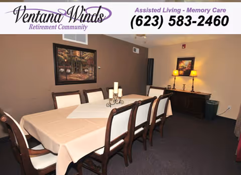 A dining room with a long rectangular table covered with a beige tablecloth and a white runner. The table is surrounded by eight wooden chairs with white cushions. On the table is a decorative candle holder with three candles. The room has brown walls, a framed painting, a sideboard with two lit lamps, and a small trash bin in the corner.