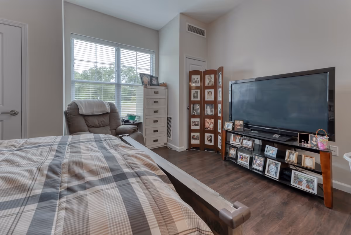 A senior living bedroom featuring a bed with a plaid comforter, a recliner chair with a white cloth draped over the back, a tall white dresser, a large flat-screen TV on a stand filled with framed photos, and a wooden folding screen displaying black and white portraits. The room has a large window with blinds and wood flooring.