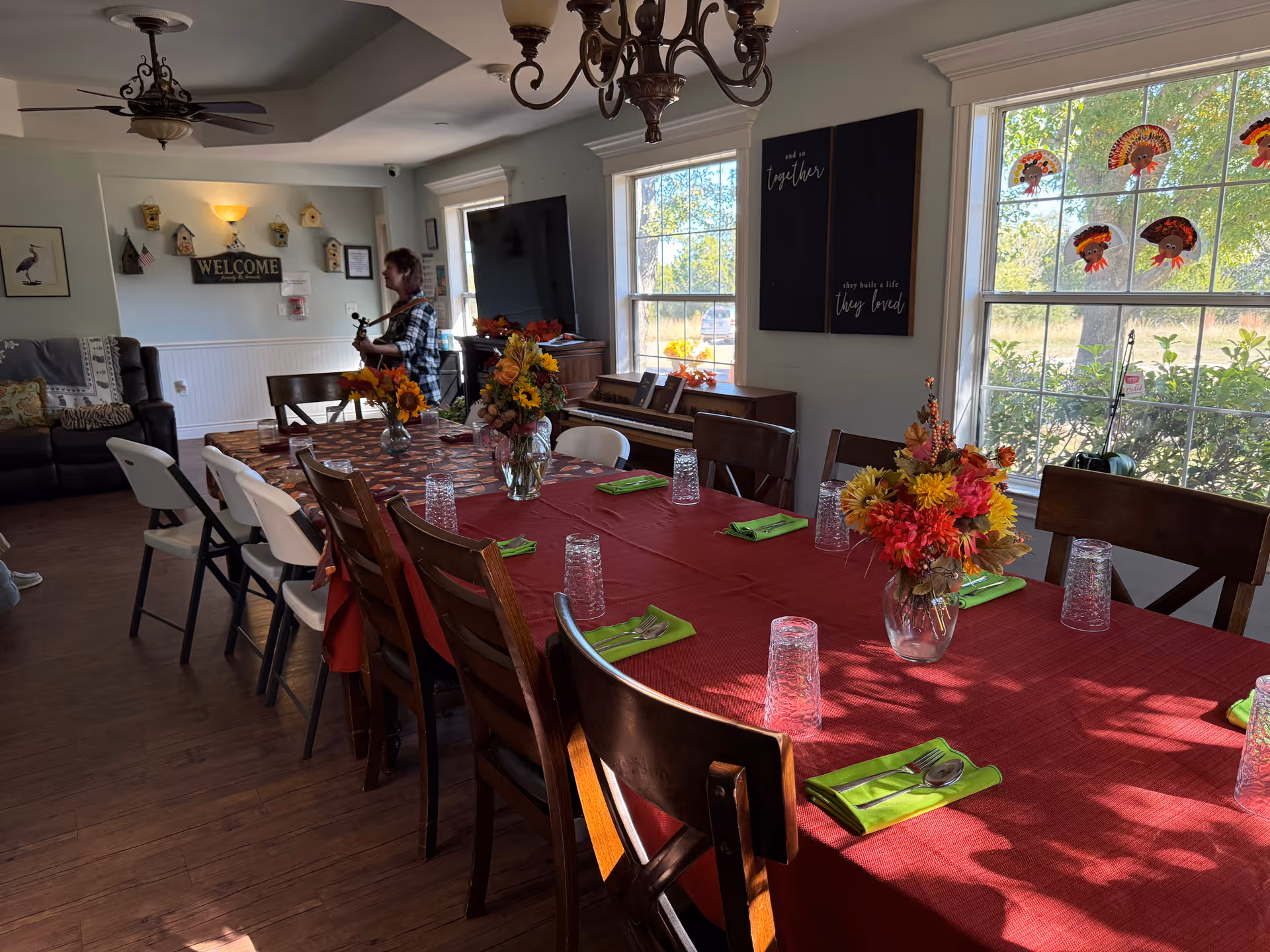 A dining room with a long table covered by a red tablecloth, set with green napkins, silverware, and clear drinking glasses. There are three vases with colorful flower arrangements on the table. The room has wooden floors, large windows with fall-themed turkey decorations, a piano, a television, and a person standing near the back wall playing a guitar. The wall has a welcome sign and birdhouse decorations.
