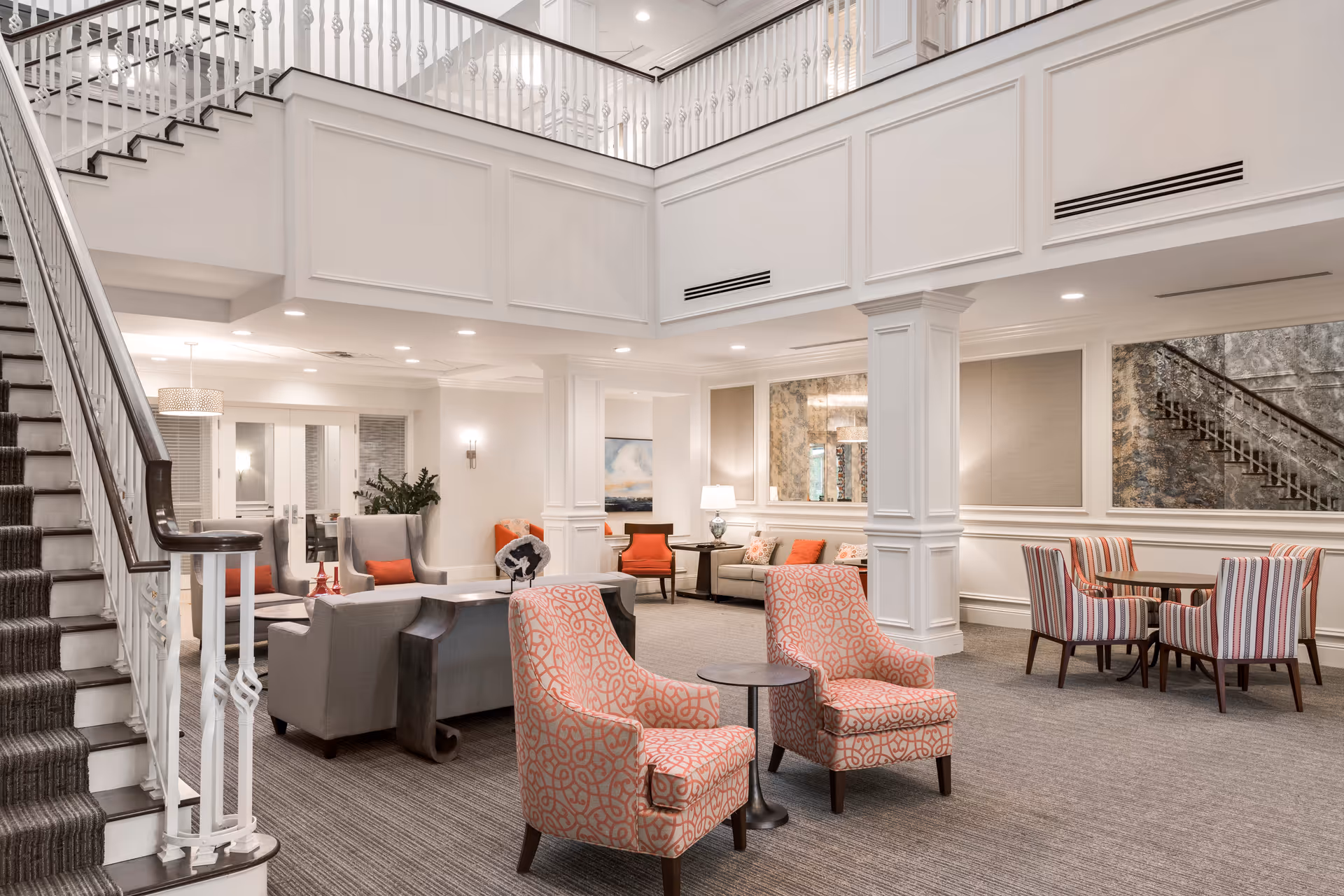 A spacious and elegant senior living facility common area with a staircase on the left, multiple seating arrangements including patterned armchairs, sofas, and small tables, white paneled walls, and soft lighting creating a welcoming atmosphere.