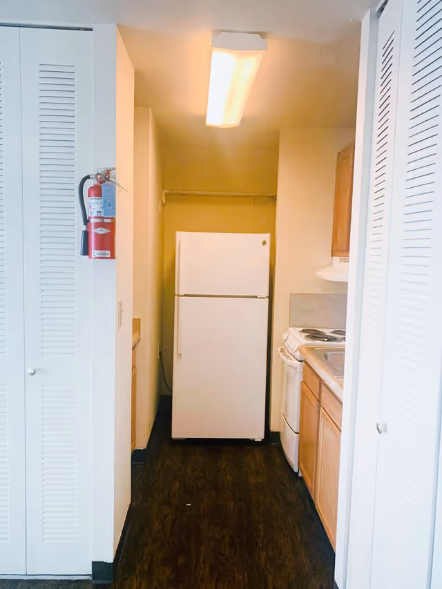 A narrow kitchen area with a white refrigerator at the end, wooden cabinets on the right, a stove and sink visible, white louvered doors on both sides, and a fire extinguisher mounted on the left wall.
