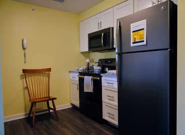 A small kitchen area with white cabinets, a black refrigerator, black stove with a striped towel hanging on the handle, and a microwave above the stove. There is a wooden chair against a yellow wall with a wall-mounted phone. The floor has dark wood-style flooring.