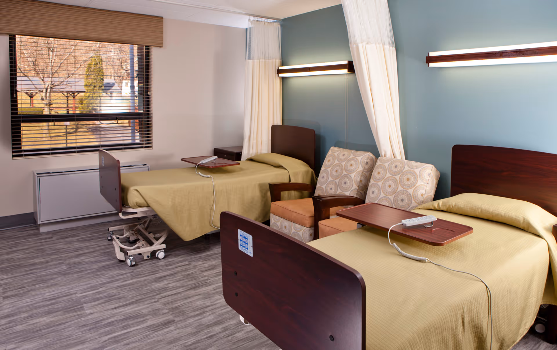A senior living facility room with two hospital-style beds covered in light green bedspreads. Each bed has a wooden headboard and a small overbed table with a remote control. Between the beds are two cushioned armchairs with circular patterned upholstery. The room has light blue and beige walls, a large window with blinds, and wood-look flooring.