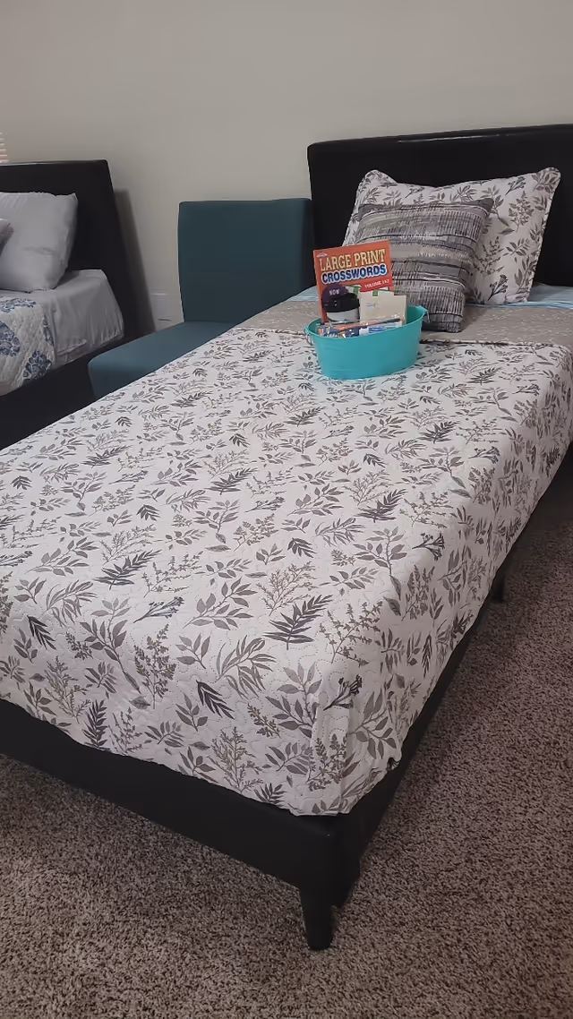 A bedroom with a twin bed covered in a floral bedspread, decorative pillows and a teal basket holding a large-print crossword book and toiletries, with another bed partially visible.