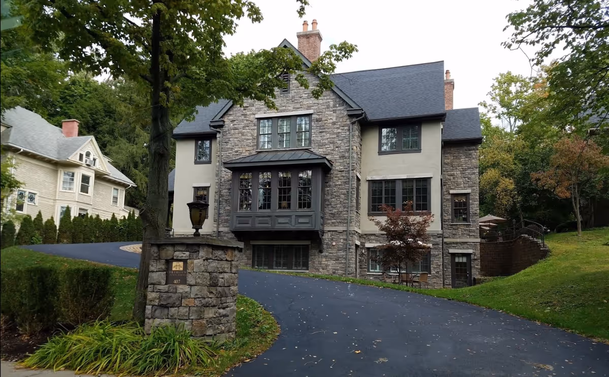 Front exterior view of a large Tudor-style stone and stucco building with multiple windows and chimneys, surrounded by trees and greenery. A curved driveway leads up to the building, with a stone pillar displaying a sign that reads 'Bridges Cornell Heights The Tudor 407'.