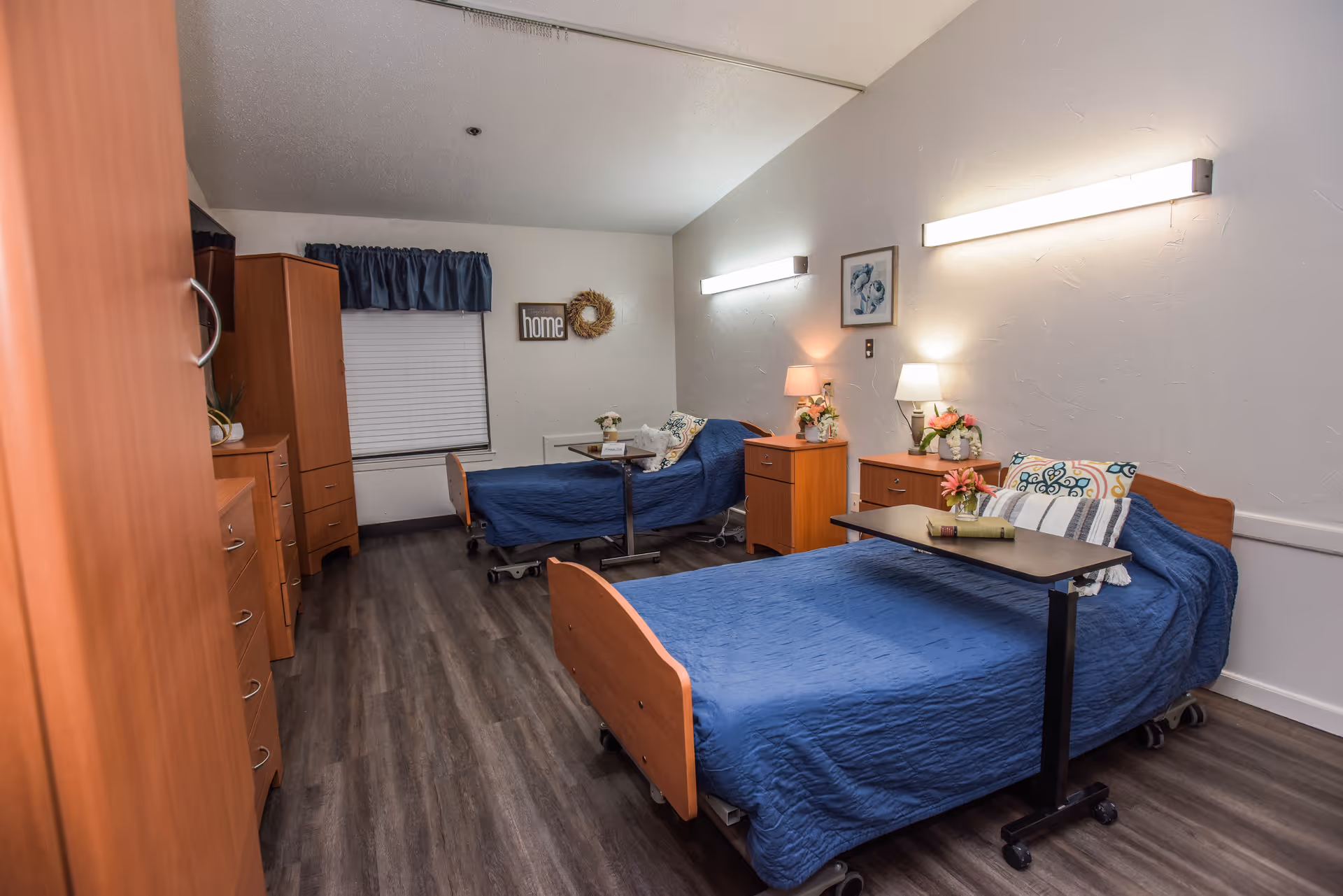 Shared senior living bedroom with two adjustable beds dressed in blue bedspreads, bedside dressers, lamps, and over-bed tables.