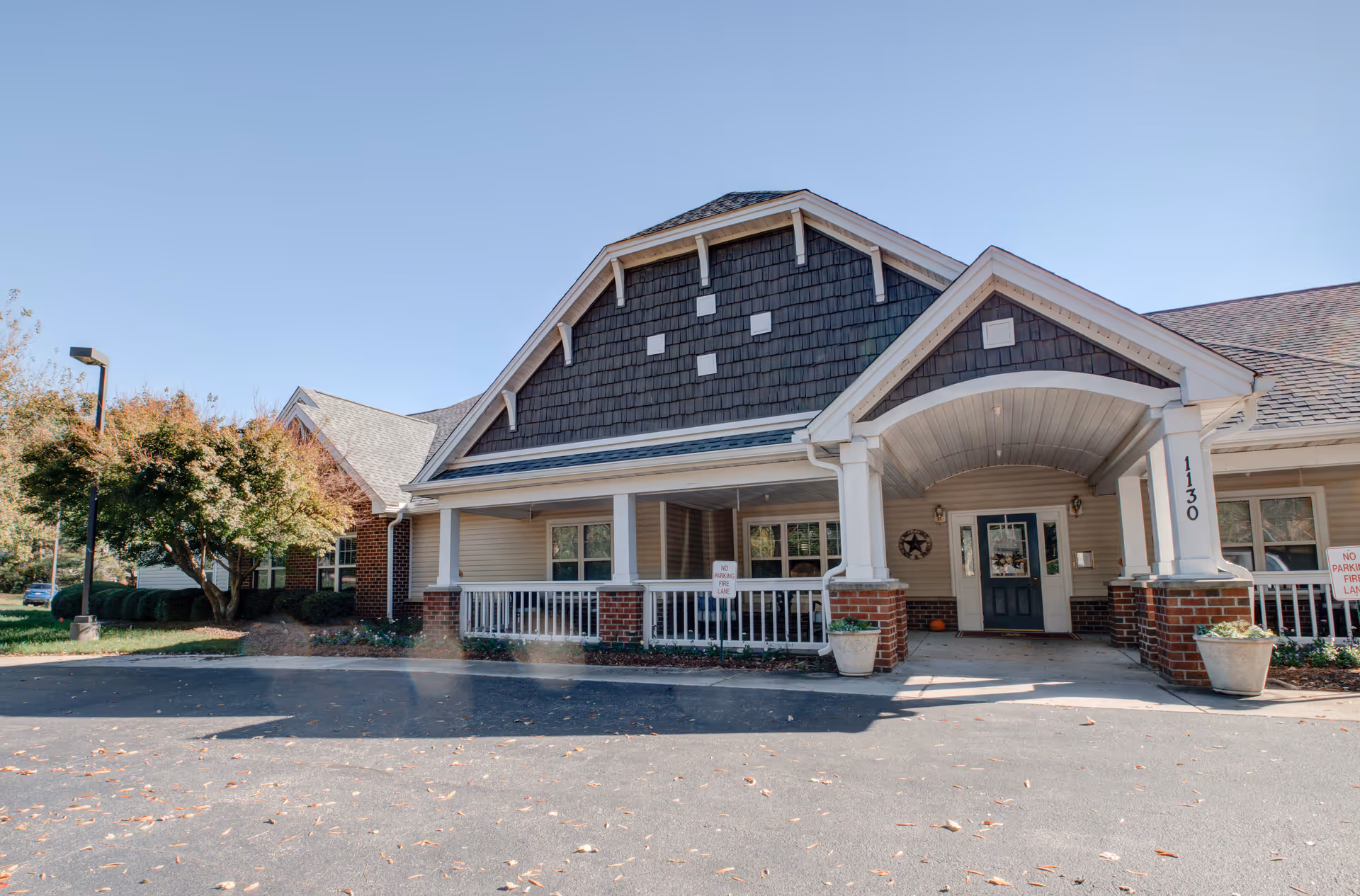 Front exterior view of Falls River Court Memory Care facility showing a covered entrance with white columns and brick accents, a dark shingled gable roof, and a driveway in front. There are some trees and landscaping on the left side under a clear blue sky.