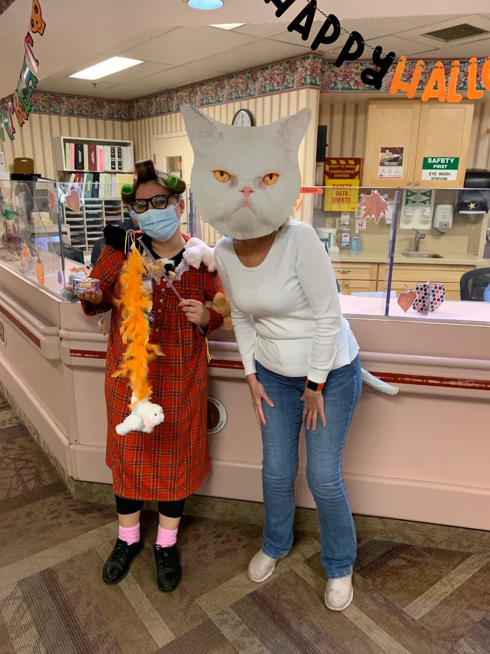 Two women standing in a reception area decorated for Halloween. One woman is wearing a red plaid dress, pink socks, black shoes, a face mask, glasses, and hair rollers, holding Halloween-themed decorations. The other woman is wearing a white long-sleeve shirt, blue jeans, and beige shoes, with a large cat face mask covering her head. The background shows a counter with a clear protective barrier, office supplies, and Halloween decorations hanging above.