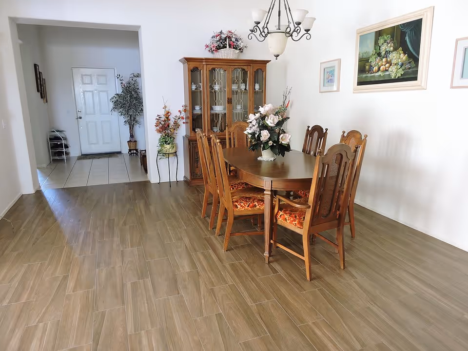 A dining room with a wooden dining table and six matching chairs with floral cushions. A flower arrangement is placed on the table. Behind the table is a wooden china cabinet with glass doors displaying dishes. The walls are decorated with framed paintings and there is a chandelier hanging from the ceiling. The floor is covered with wood-like tiles, and there is a doorway leading to a tiled entryway with a white door and a potted plant.