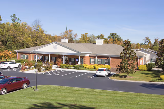 Exterior view of a single-story brick building with a covered entrance, surrounded by a parking lot with several cars and landscaped greenery including trees and bushes under a clear sky.