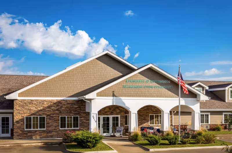 Front exterior of a senior living community building with a covered arched entrance, American flag, and landscaped driveway.