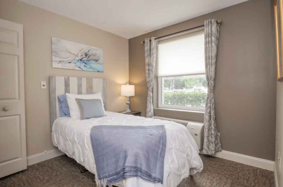 A cozy bedroom with a single bed made with white bedding and a light blue throw blanket. The bed has a wooden headboard with a striped pattern. There is a bedside table with a lamp, a window with patterned curtains, and a piece of abstract art hanging on the wall above the bed.