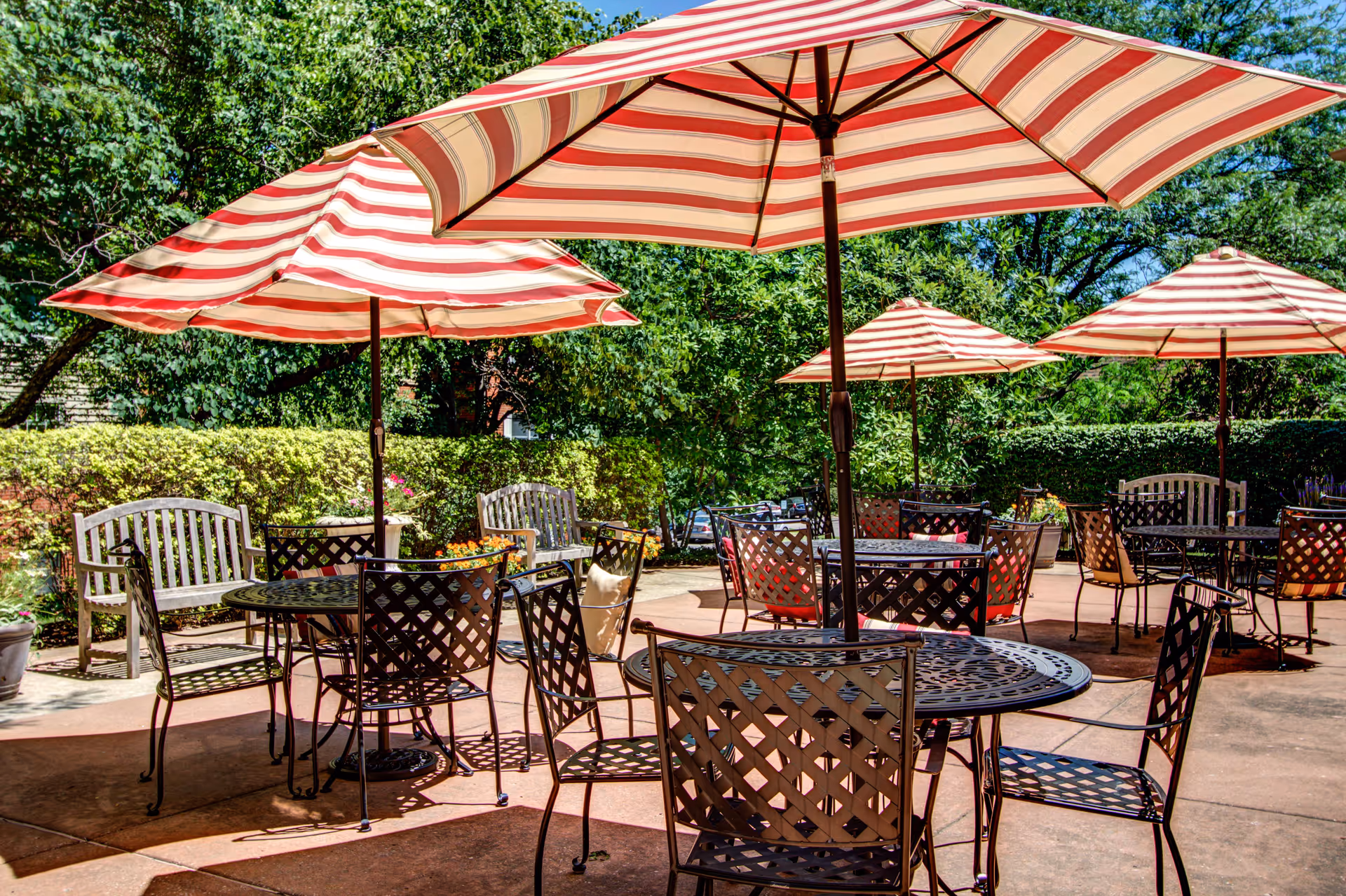 Outdoor patio with metal tables and chairs under red-and-white striped umbrellas surrounded by greenery.