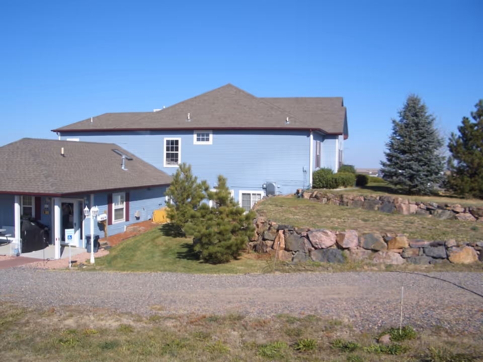 Light blue two-story house with a gravel driveway, stone retaining wall, and small trees under a clear blue sky.