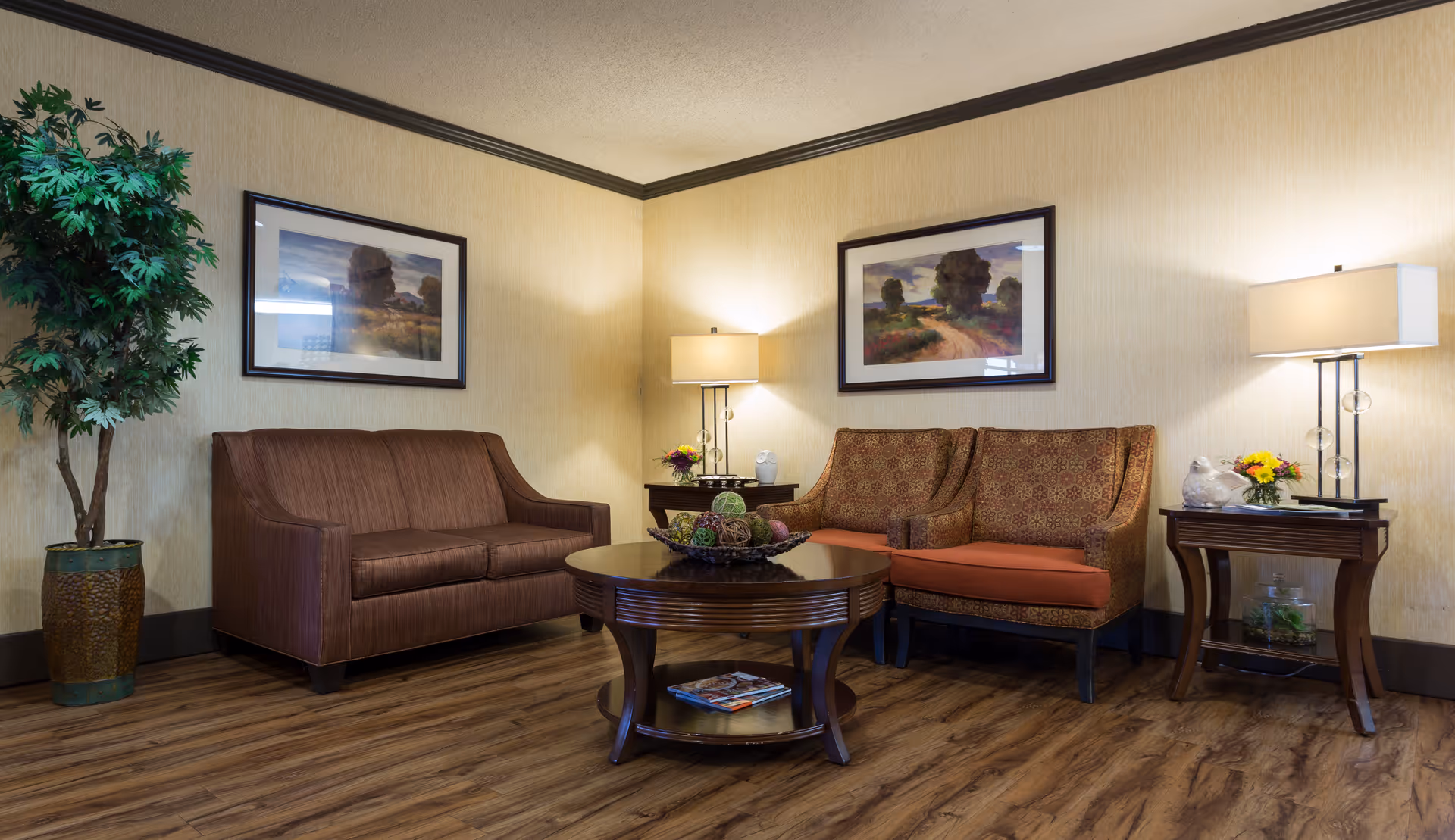 A cozy sitting area with a brown sofa, two patterned armchairs with orange cushions, a round wooden coffee table with decorative items, two side tables with lamps, a potted plant, and framed landscape paintings on the walls.