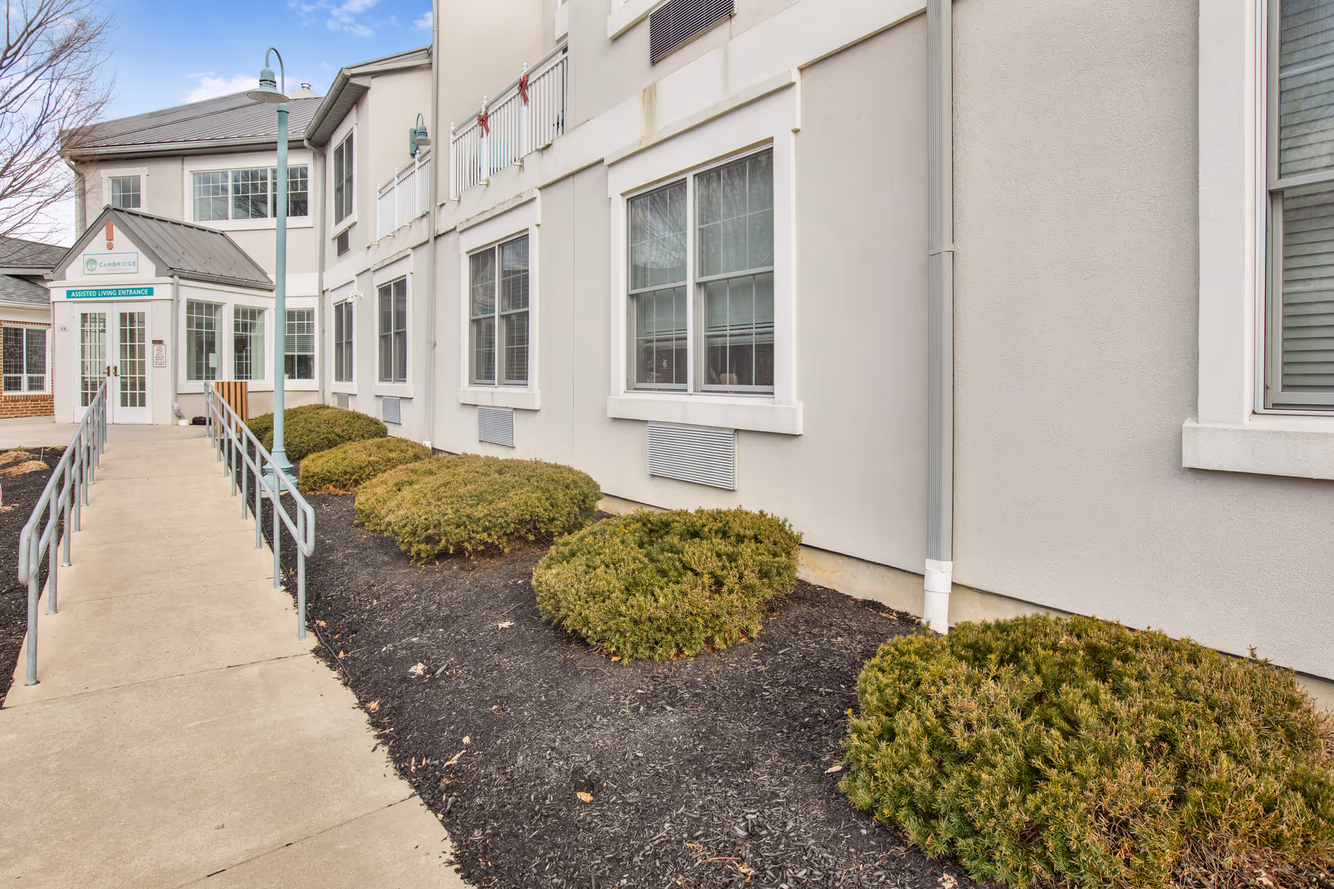 Exterior view of a senior living facility showing a concrete walkway with handrails leading to a glass entrance door labeled 'Assisted Living Entrance'. The building is light-colored with multiple windows and trimmed bushes along the walkway.